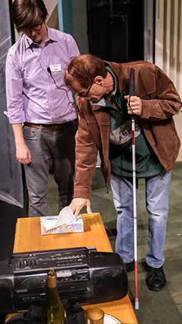 A man in a brown jacket with a visual impairment is accompanied by a man in a purple shirt as he touches props on stage from the production of 